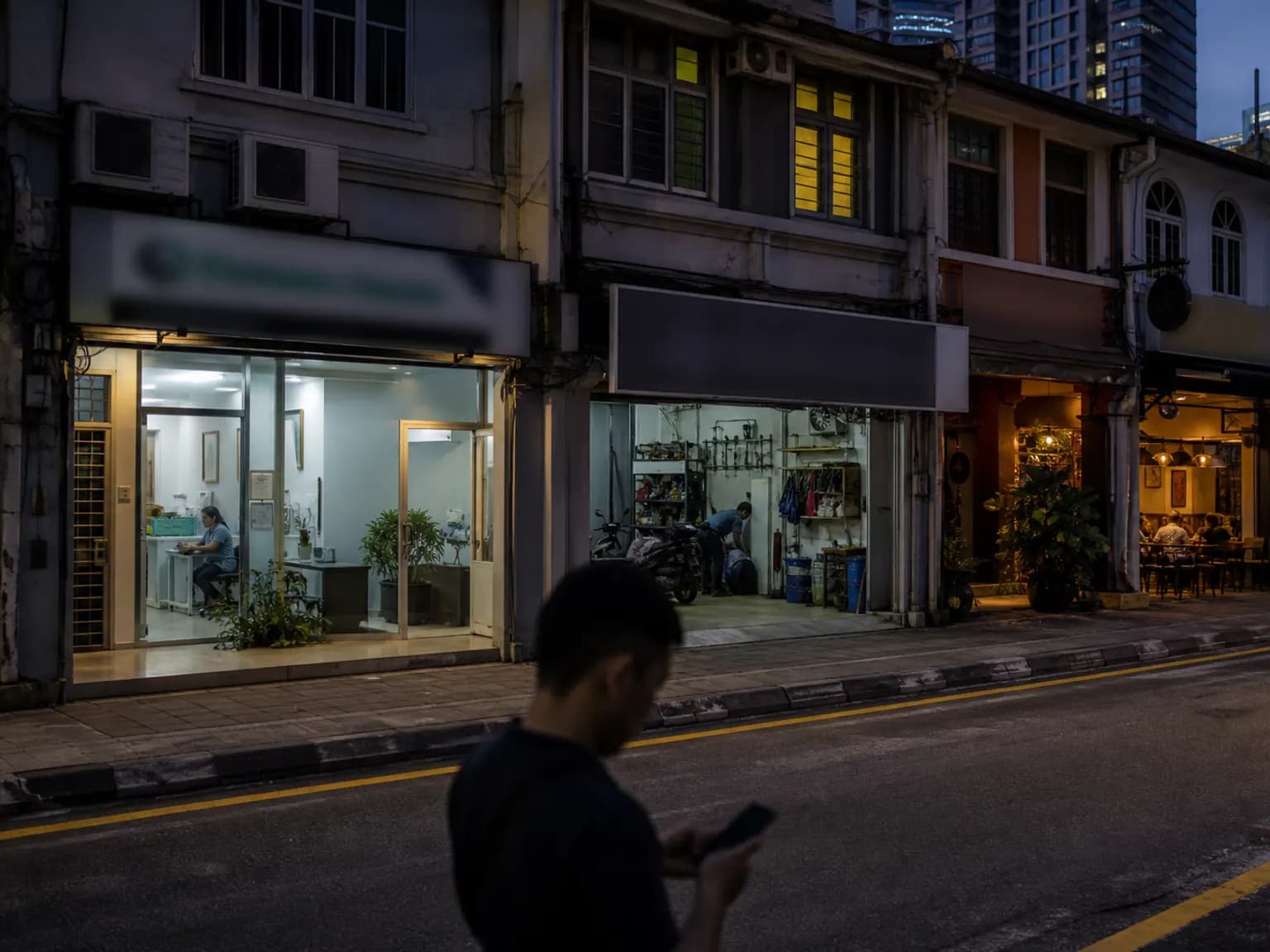 Kuala Lumpur shophouse street at dusk with a clinic, a workshop and a cafe open, and a passer-by checking his phone.