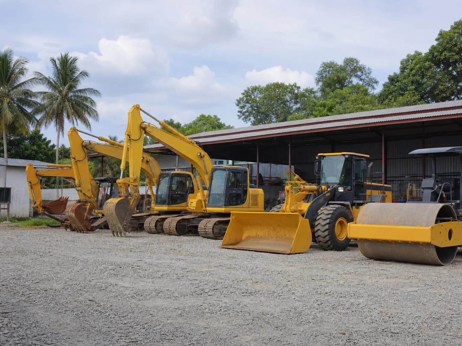 Excavators, a wheel loader, and a road roller parked in a row in a gravel equipment yard.