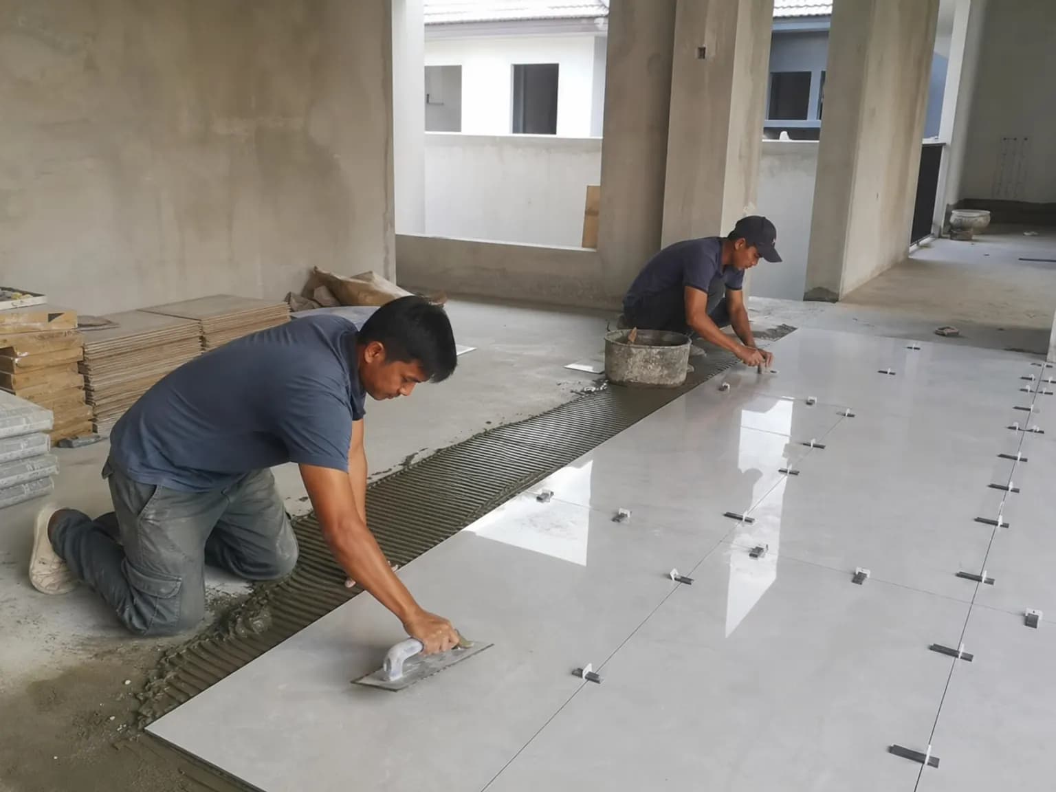 Renovation crew laying large-format floor tiles in an open living area, with bags of tile adhesive nearby.